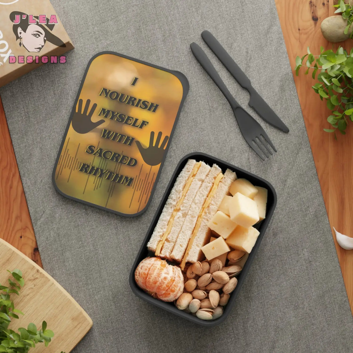 Decorative bento box with cheese, crackers, nuts, and fruit on a gray surface with a decorative mat and cutlery.